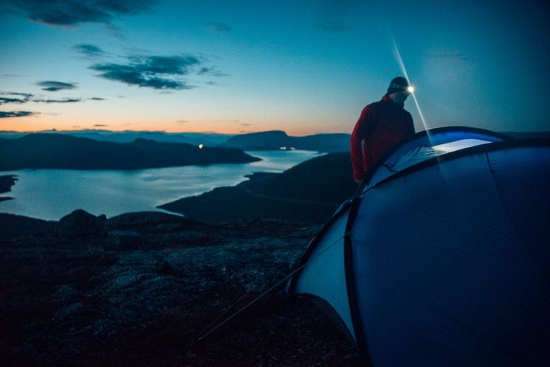 Een man met een hoofdlamp die voor zijn tent staat. De tent is opgezet bovenop een heuvel met uitzicht op een meer.