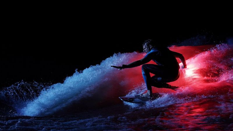 Een surfer die 's nachts op een golf surft, verlicht door levendige rode en blauwe lichten die een opvallend contrast creëren met de donkere achtergrond.