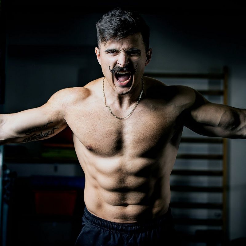 A muscular, shirtless athlete with a handlebar moustache grimaces with effort as he grips on to gymnastic rings. 