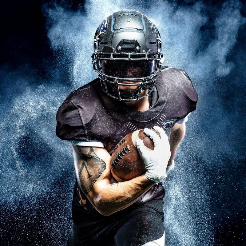 Игрок в американский футбол держит мяч одной рукой и словно бежит в сторону камеры в окружении белого дыма и пыли.