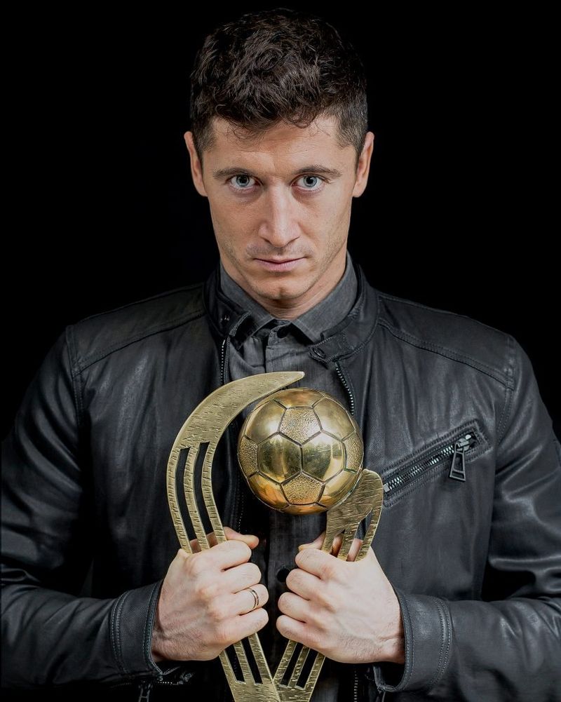 Профессиональный футболист Роберт Левандовский в кожаной куртке и темно-серой футболке держит перед собой золотой кубок.