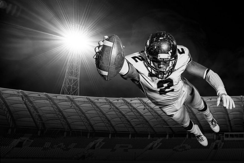 Черно-белое изображение игрока в американский футбол, ныряющего с мячом в вытянутой руке под светом ярких прожекторов стадиона.