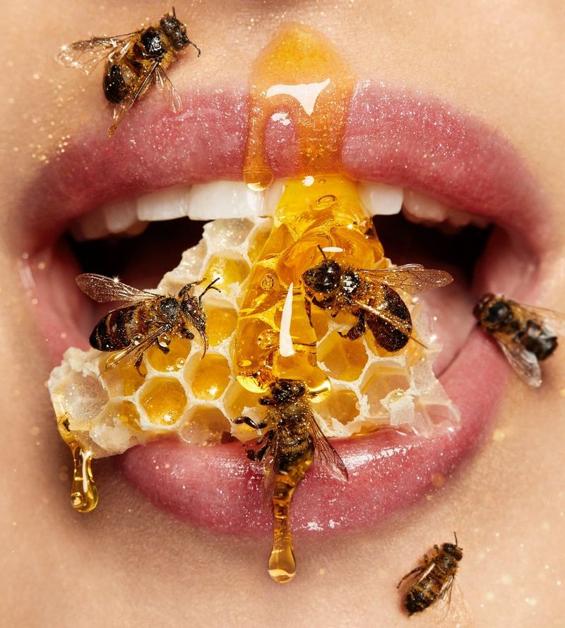 A close-up of a woman's mouth, holding a piece of dripping honeycomb between her teeth and bottom lip, with several bees crawling over the honey and her skin, by macro photographer Tina Eisen.