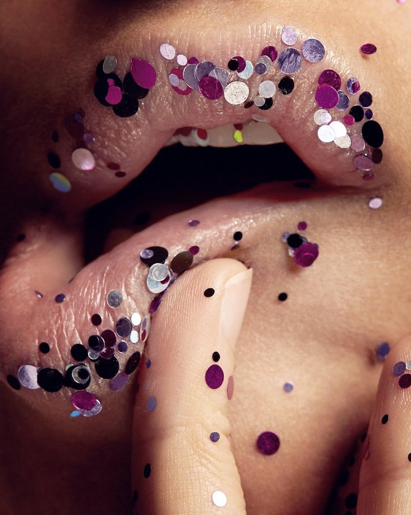 A close-up of a woman's mouth, lightly covered in purple glitter and with the bottom lip being pushed to one side by a finger, in a macro photograph by Tina Eisen.