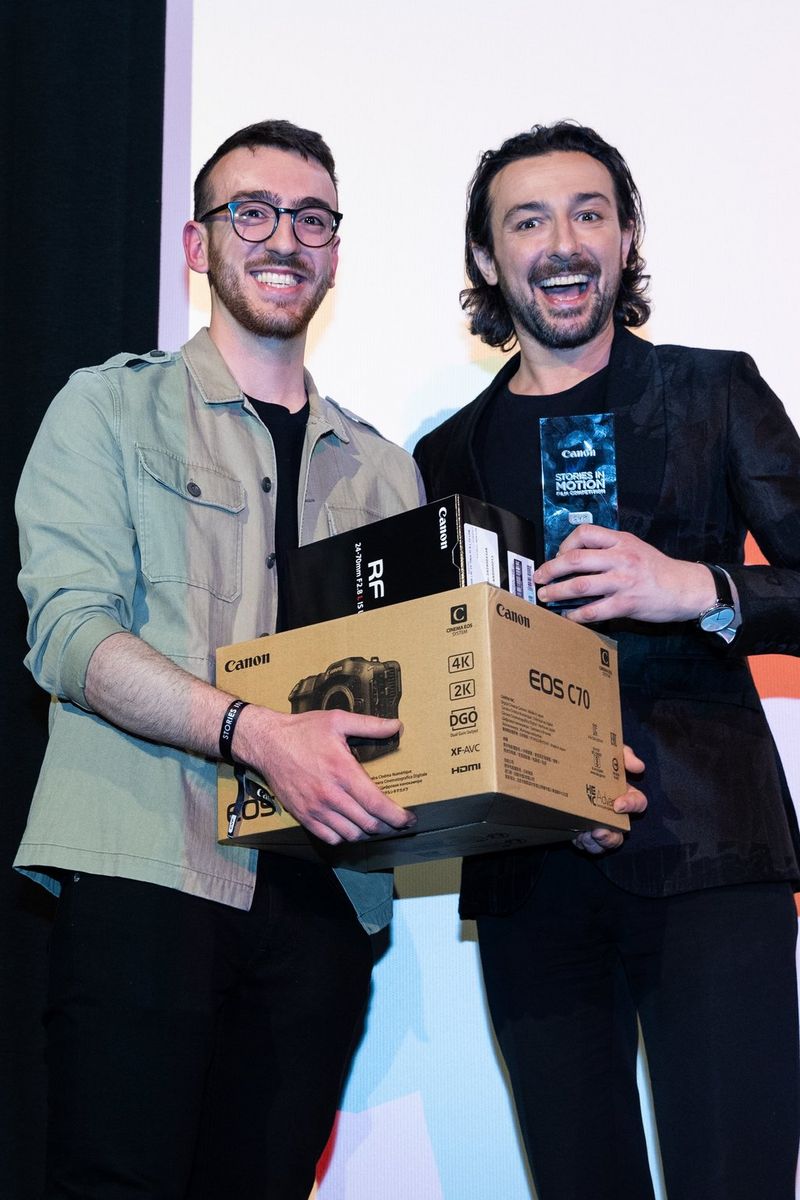 Young filmmaker Daniel Simpkins stands holding a pile of boxed Canon equipment, standing next to host Alex Zane who is presenting him with his Grand Prize award in the Canon Stories in Motion Young Filmmakers Competition.