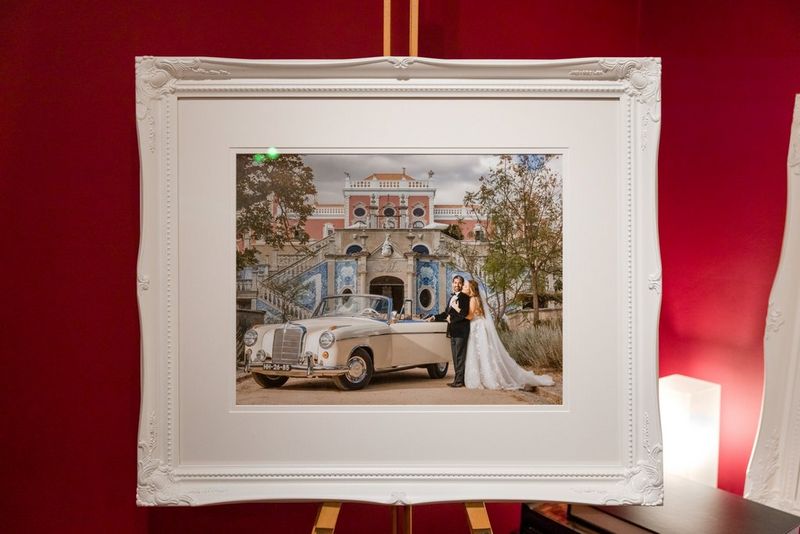 A large print of a wedding photo, showing a bride and groom next to a cream convertible Mercedes car in front of a grand building. The print is framed and standing on an easel in a red-painted room.
