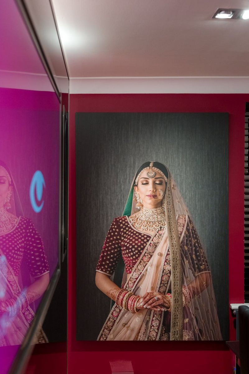 A large print of an Indian woman in a deep red wedding sari embroidered with gold, plus gold jewellery and henna patterns on her hands, hangs on a red wall. It is reflected in a plastic panel on another wall to the left of the frame.