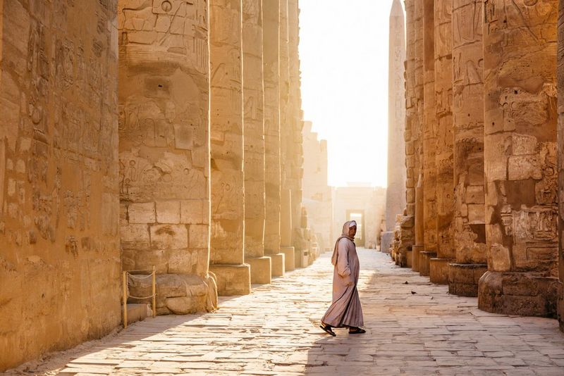 Een persoon in een lang, wit en crèmekleurig gewaad en sandalen staat tussen twee lange, gewelfde muren die tot hoog in de lucht reiken.