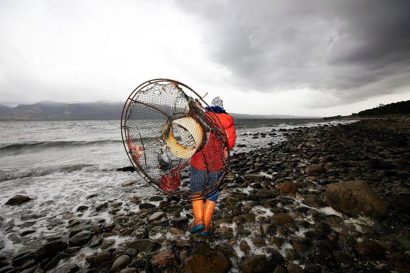 Een man in een regenjas loopt weg van de camera op een nat strand vol stenen. Hij draagt een groot, rond visnet.