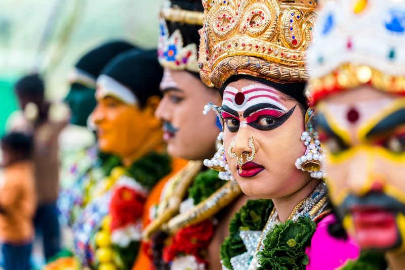Een rij mensen met felgekleurde kleding, decoratieve hoofdbedekking en make-up. Twee gezichten zijn naar de camera gericht, één ervan is scherp in focus.