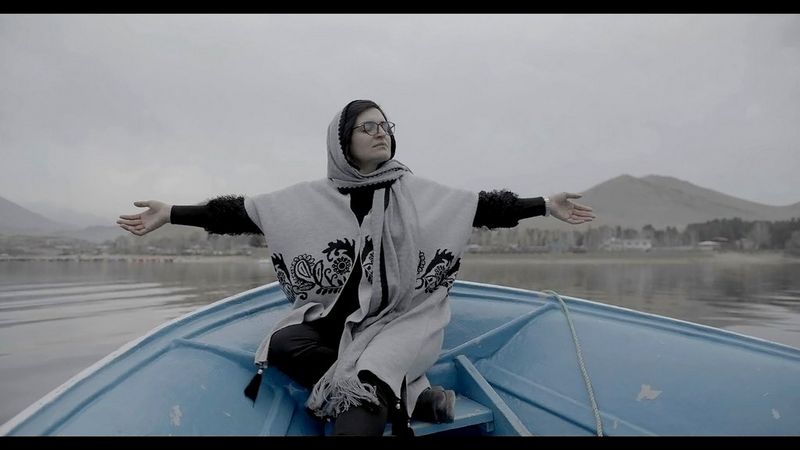 A still from In Her Hands showing Zarifa Ghafari sitting in a blue boat with her arms spread wide and her eyes closed, filmed by Marcel Mettelsiefen.