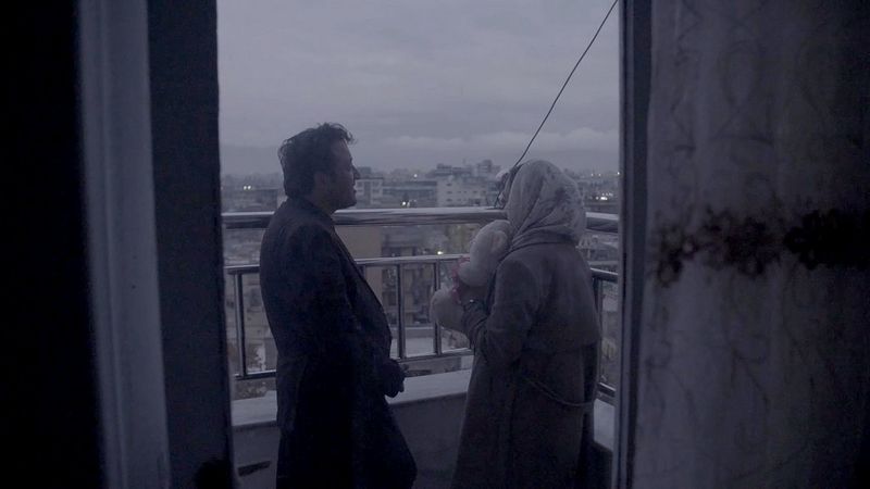 A still from In Her Hands showing a man and Zarifa Ghafari standing in a balcony overlooking a city, while Zarifa holds a stuffed toy in her hands, filmed by Marcel Mettelsiefen.