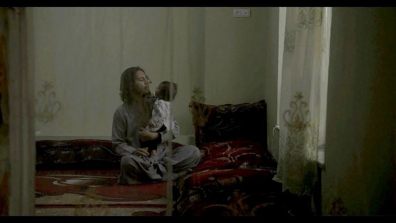 A still from In Her Hands showing a man sitting on the carpet and holding a child in his arms, filmed through gauze curtains by Marcel Mettelsiefen.