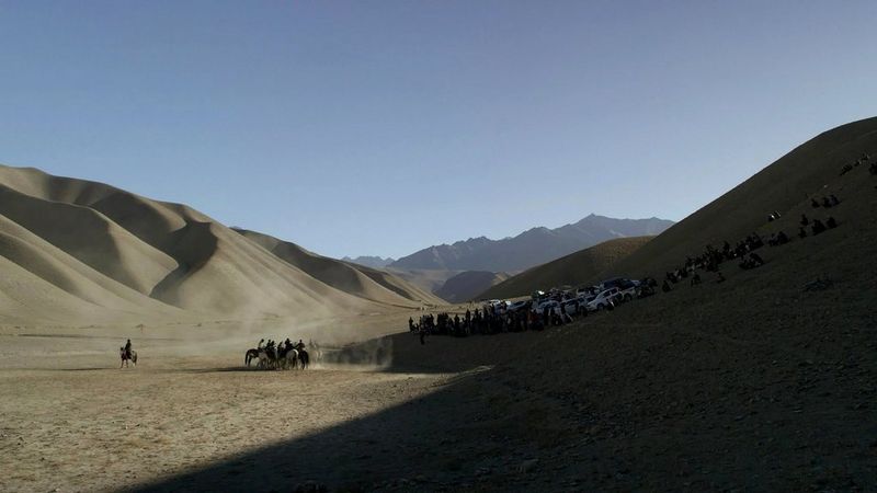 A still from In Her Hands showing a desert landscape in Afghanistan, with men on horses gathered to the left of the frame and people sitting in the shadows to the right of the frame, filmed by Marcel Mettelsiefen.