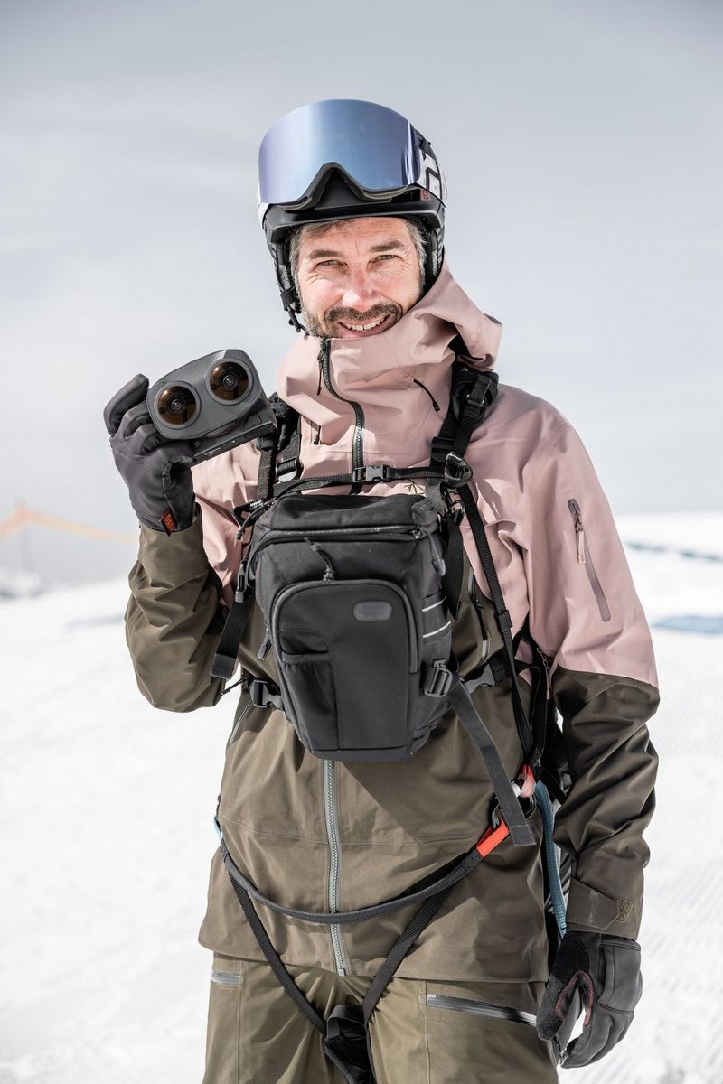 Un uomo con abbigliamento outdoor, casco e una borsa fotografica sul petto, tiene in mano una fotocamera con obiettivo Dual Fisheye.