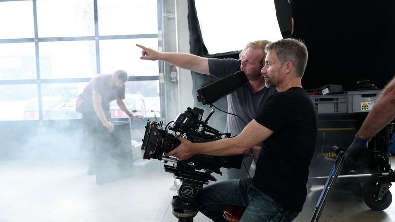 Un uomo inclina una fotocamera Canon verso l'alto su un set cinematografico polveroso, mentre un secondo uomo gli indica qualcosa in lontananza.