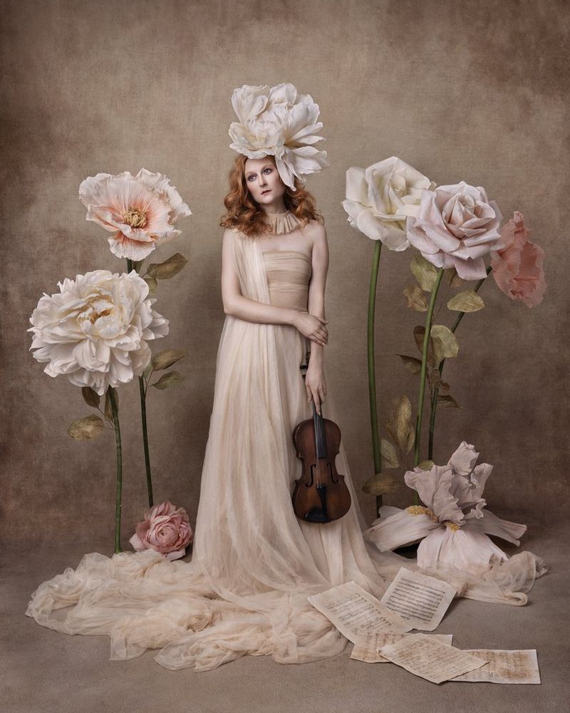 Een vrouw in zijden stof houdt een viool aan haar zijde en er ligt bladmuziek op de grond. Een grote bloem op haar hoofd is dezelfde als enorme bloemen naast haar. Gefotografeerd met een Canon EOS R5 en een RF 24-105mm F4L IS USM-objectief.