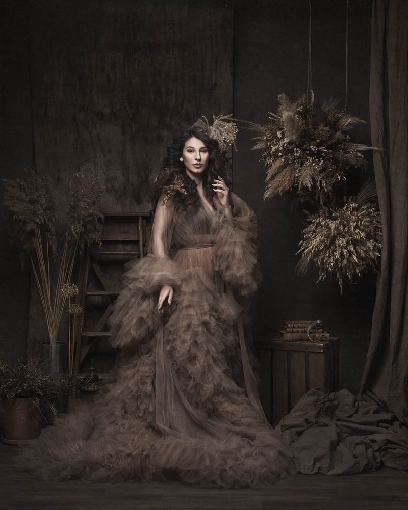Een portret van een vrouw met donker haar die een lange bruine jurk draagt met veel lagen ruches. Ze staat in een ruimte met een houten ladder, gedroogde bloemstukken en gedrapeerde stof. Gefotografeerd met een Canon EOS R5 en een RF 24-105mm F4L IS USM-objectief.