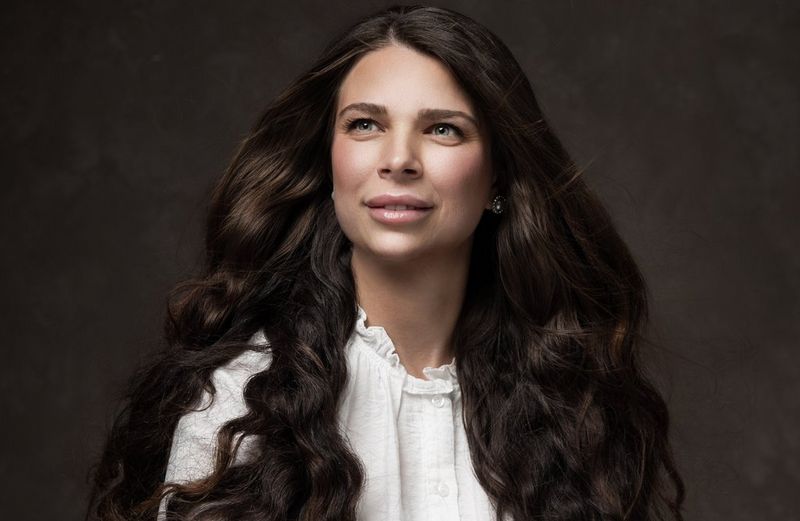 Een sereen portret door Martina Wärenfeldt van een vrouw met lang, golvend bruin haar en blauwe ogen op een donkere achtergrond. Gefotografeerd met een Canon EOS R5 en een RF 24-105mm F4L IS USM-objectief.