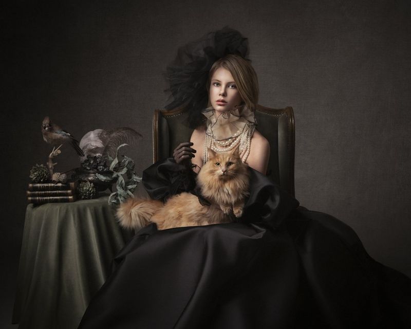 Een vrouw met een donkere zijden rok zit met een langharige kat op haar schoot. Naast haar staat een tafel met planten, veren, boeken en een opgezette vogel. Gefotografeerd met een Canon EOS R5 en een RF 24-105mm F4L IS USM-objectief.