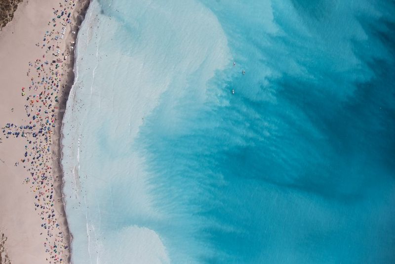 An aerial view of the shoreline, with hundreds of people enjoying the sandy beach and azure-blue ocean waters.