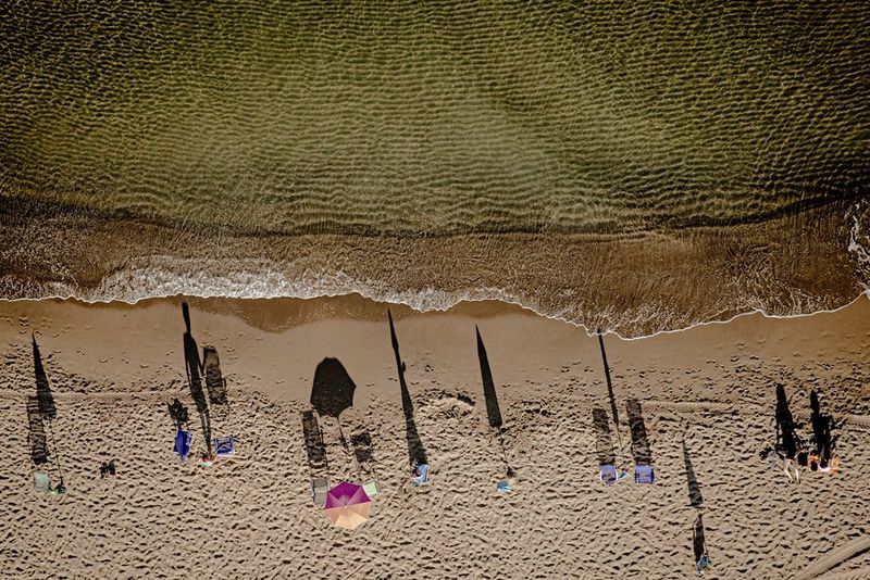An aerial image of people and umbrellas on a beach casting long shadows towards the waves breaking on the shore.