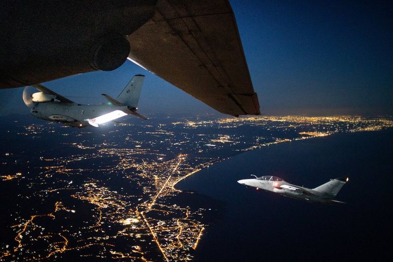 In the sky above the lights of a city at night, an aircraft is illuminated by the light from the open cargo bay of a large plane.