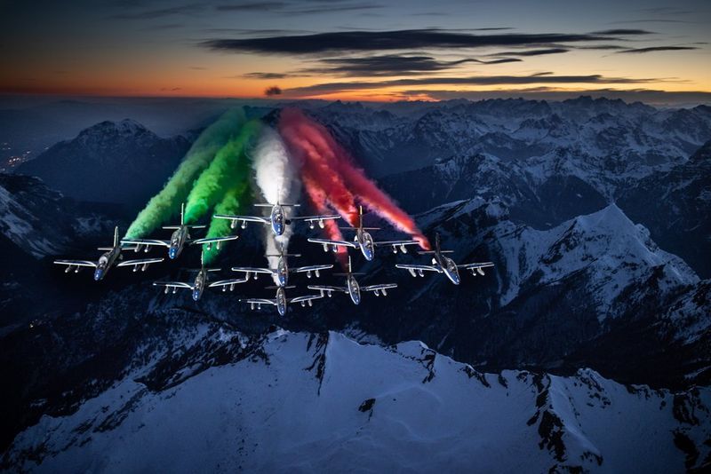An aerial image of Italy's Air Force aerobatic display team flying above a snow-covered mountain range in the twilight, trailing green, white and red coloured smoke.