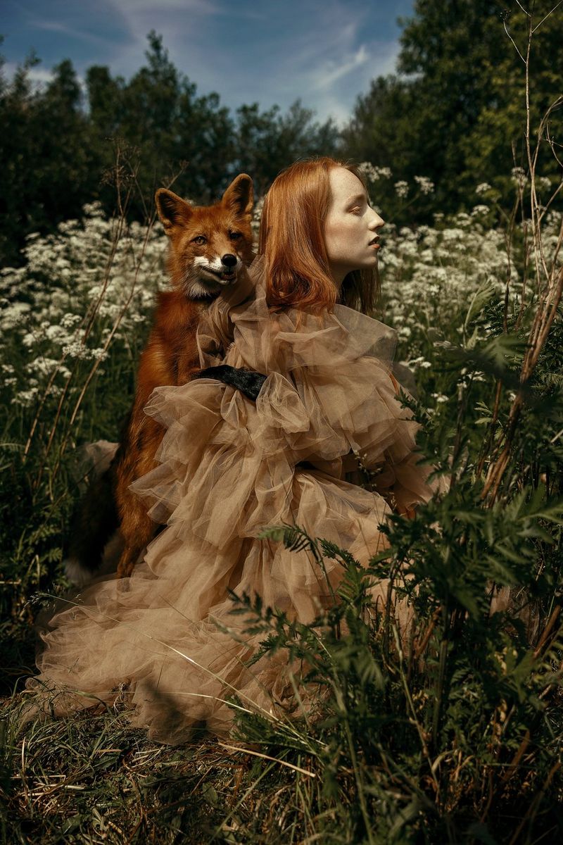 Une jeune femme rousse portant un renard sur son dos est debout dans un champ. On aperçoit des arbres au loin. Photo prise par Menna Hossam avec un Canon EOS R5.