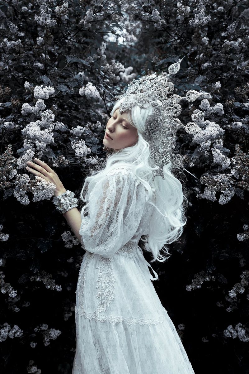 Une femme vêtue d'une robe blanche et d'une couronne caresse des fleurs blanches avec sa main gauche. Photo inspirée par le Monde de Narnia et prise par Menna Hossam avec un Canon EOS R5.