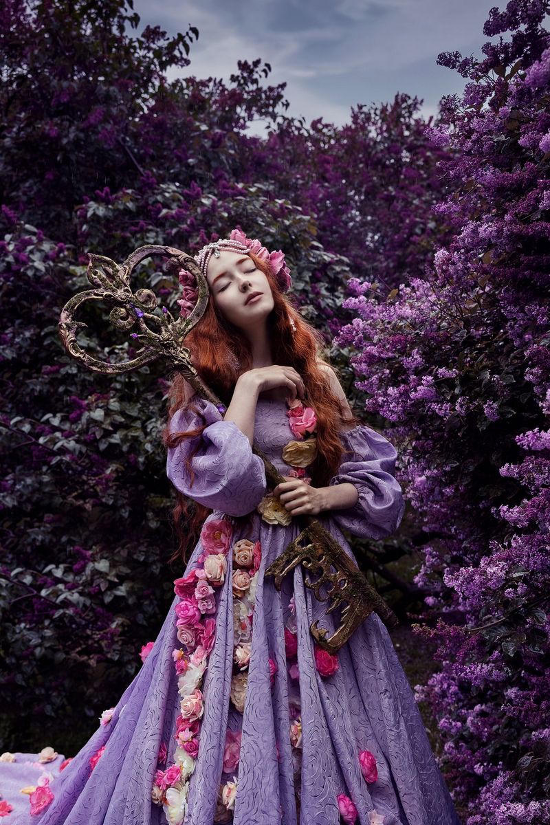 Une femme portant une somptueuse robe violette recouverte de fleurs roses et enlaçant une énorme clé se tient devant un feuillage luxuriant. Photo prise par Menna Hossam avec un Canon EOS R5.