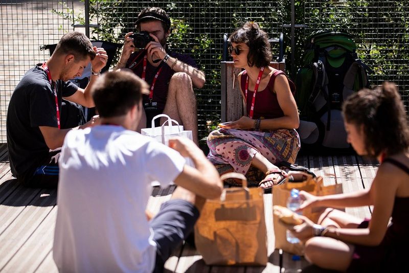 A group of people sit on wooden decking, eating packed lunches from paper bags. They all wear lanyards with red straps around their necks, and one man is holding up a Canon EOS R and looking through the viewfinder