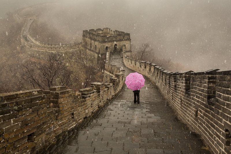 Sneeuw valt op een persoon die op de Chinese Muur staat en een opvallende roze paraplu vasthoudt. Gefotografeerd door Joel Santos.