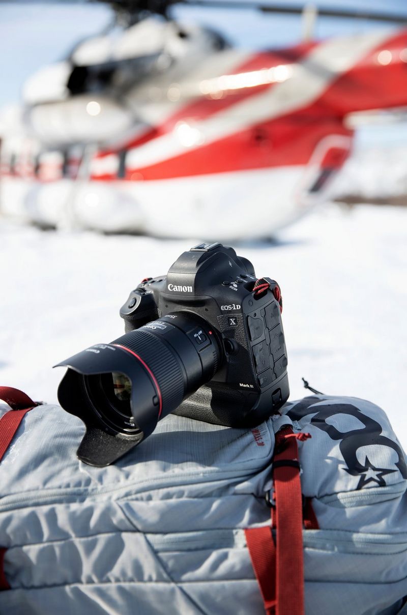 A Canon EOS-1D X Mark III perched on top of a rucksack. A helicopter can be seen on the snow-covered ground behind.