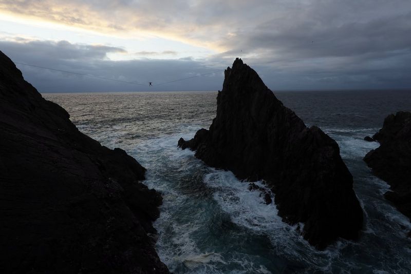 A view from some distance of a slackline walker on a line between rocky cliffs over swirling water.