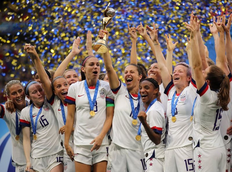 Carli Lloyd, captain of the US women's national football team, lifts a glittering gold trophy above her head as her teammates celebrate in a shower of confetti.