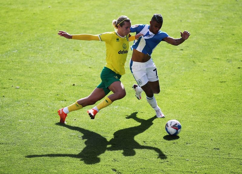 Norwich City's Todd Cantwell and Huddersfield Town's Juninho Bacuna jostle for the ball on a football pitch bathed in sunlight.
