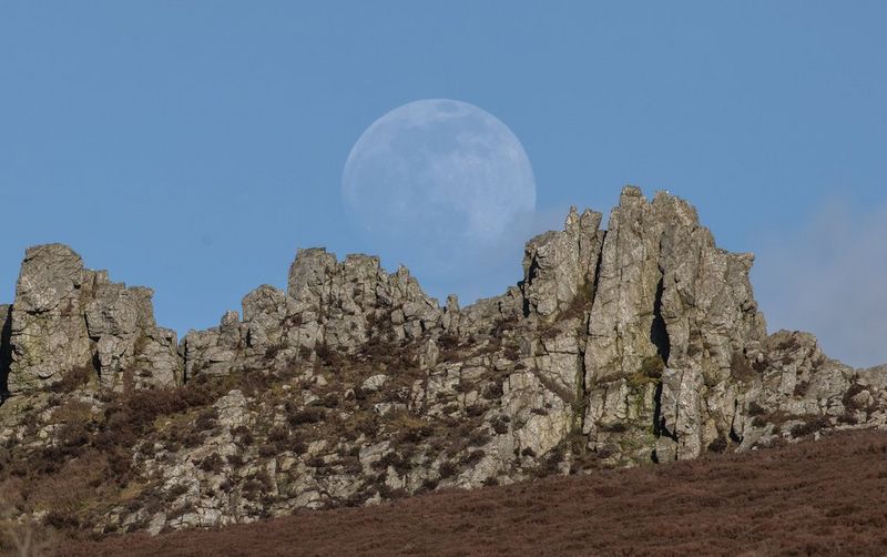 Размытая Луна поднимается над каменистым выступом холма Стиперстонс в графстве Шропшир, Великобритания. Снято Эндрю Фьюзеком Питерсом на камеру Canon EOS R5 с объективом Canon RF 600mm F4L IS USM.