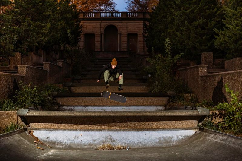 Alors que l'obscurité tombe, un skateboardeur réalise un trick sur de larges marches dans un jardin richement décoré, dans une photographie droite prise avec un appareil photo Canon EOS R6 par Josh Katz.