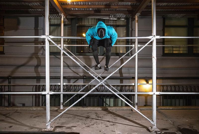 Homme assis sur un barreau d'échafaudage à plusieurs mètres au-dessus du sol, photographié en train de se balancer autour de la barre.