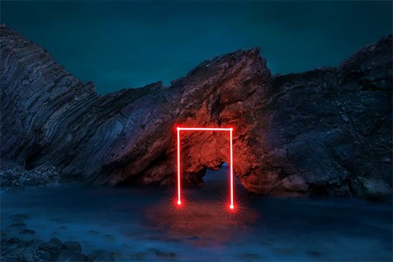 A multiple exposure image of a doorframe outlined in red light. The shape appears to emerge from the sea and is set against a rocky backdrop.