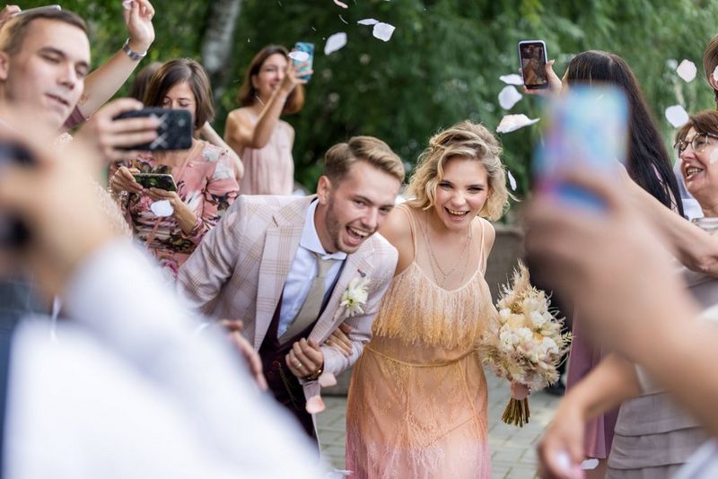 Пара улыбается и смеется среди дождя из конфетти, находясь в окружении друзей и родственников, которые снимают их на телефоны.