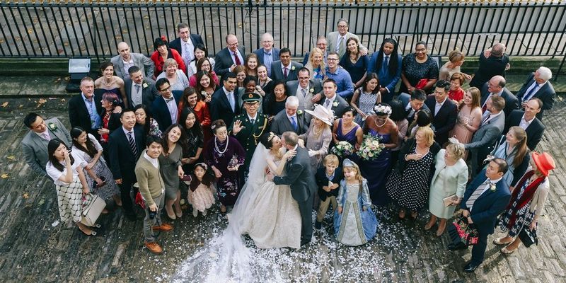 Фотограф смотрит сверху вниз на большую группу родственников и друзей, приглашенных на свадьбу. Жених и невеста целуются на переднем плане.