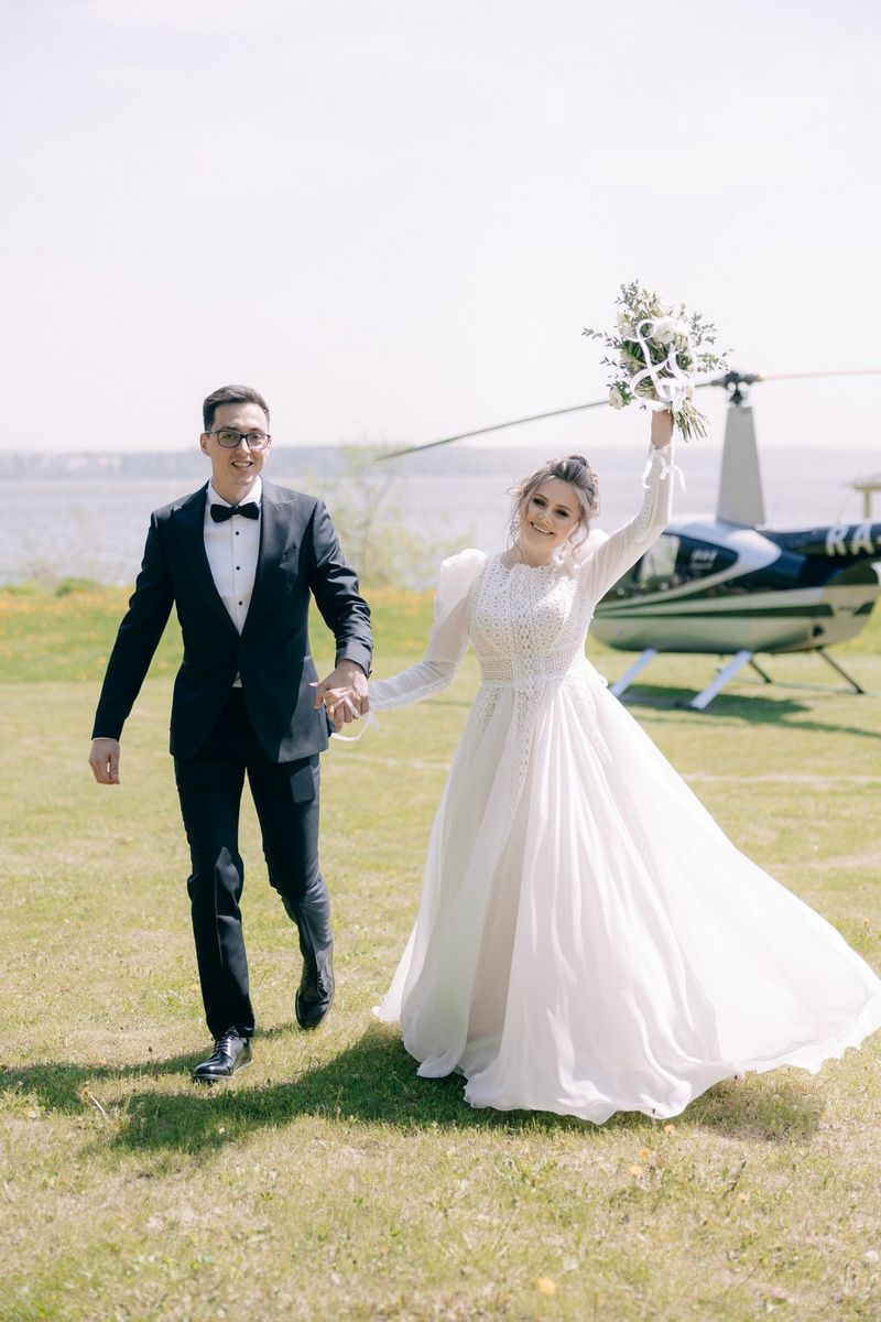 Жених и невеста держатся за руки и идут по полю вдаль от вертолета; невеста держит в вытянутой вверх руке букет.