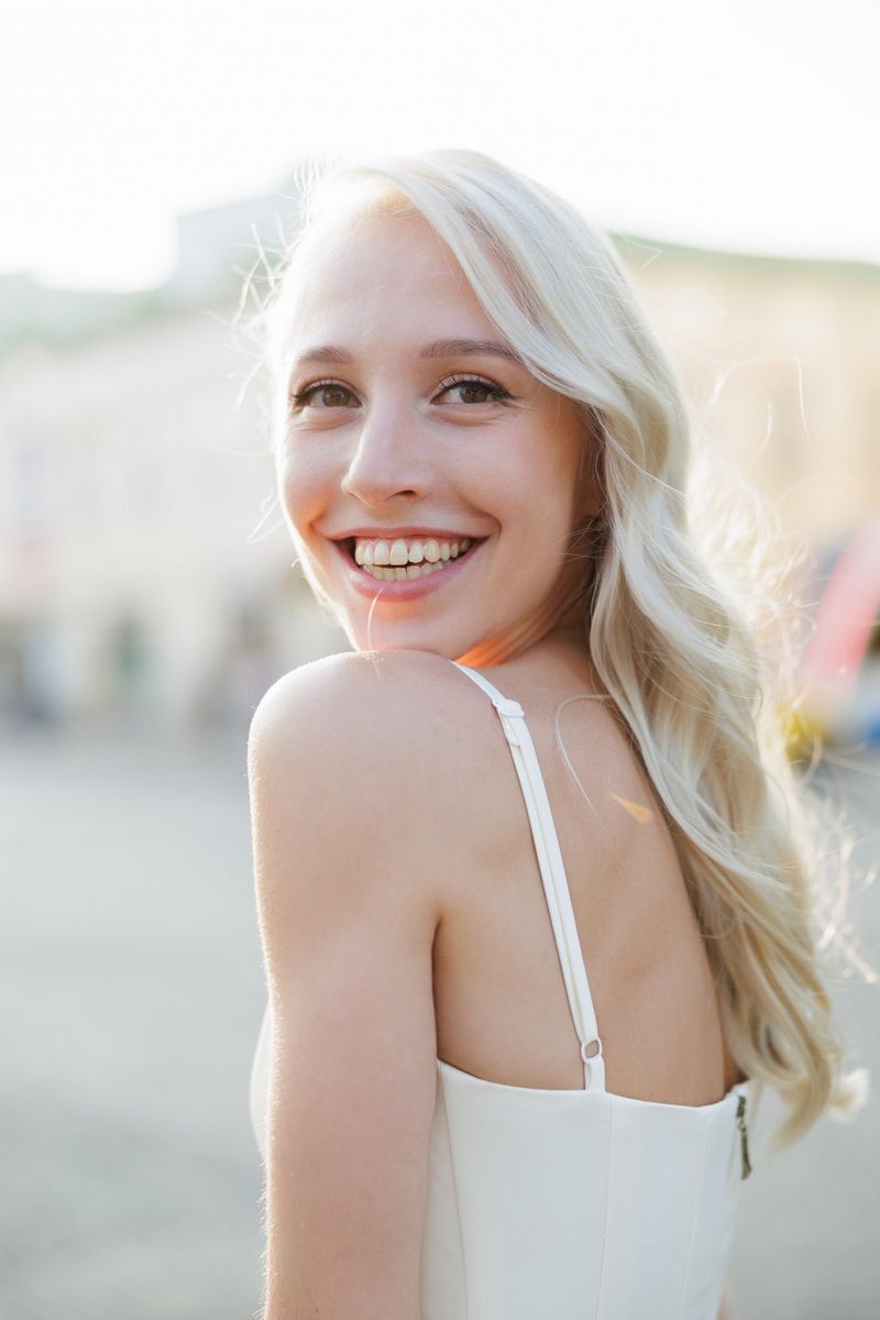Женщина в платье на бретелях смотрит через плечо в сторону камеры и улыбается.