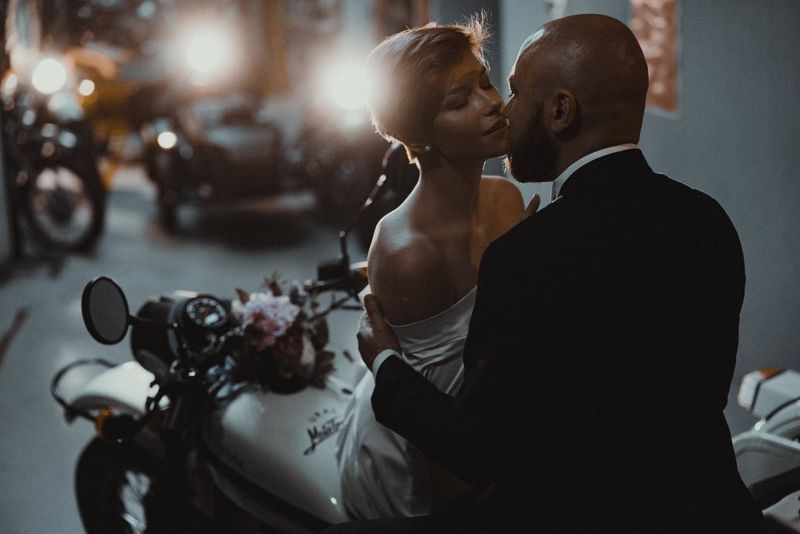 Жених с невестой целуются, сидя на мотоцикле среди других мотоциклов в гараже.