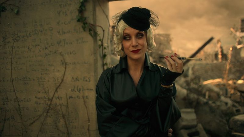 In a still from The Umbrella Academy, Kate Walsh leans against a wall with ruined buildings behind. 