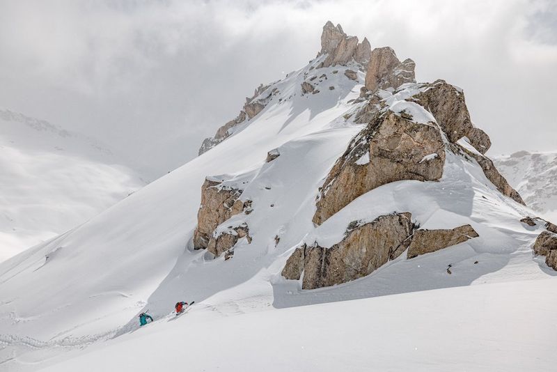 Two skiers, dwarfed by the mountain peak in front of them, climb upwards through deep snow.