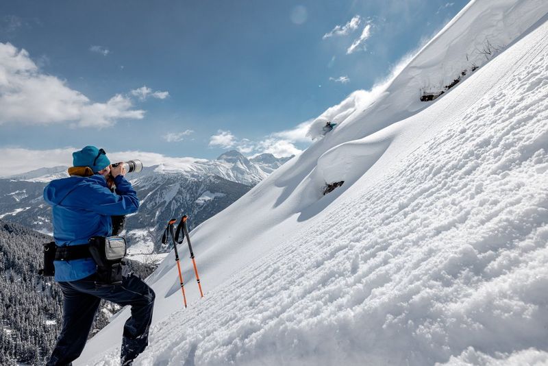 A man stands on a ski slope, filming a skier kicking up a cloud of snow.