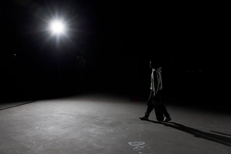 A man walking with a skateboard at night. A bright light in the upper left of the frame is creating stark shadows, obscuring his features.